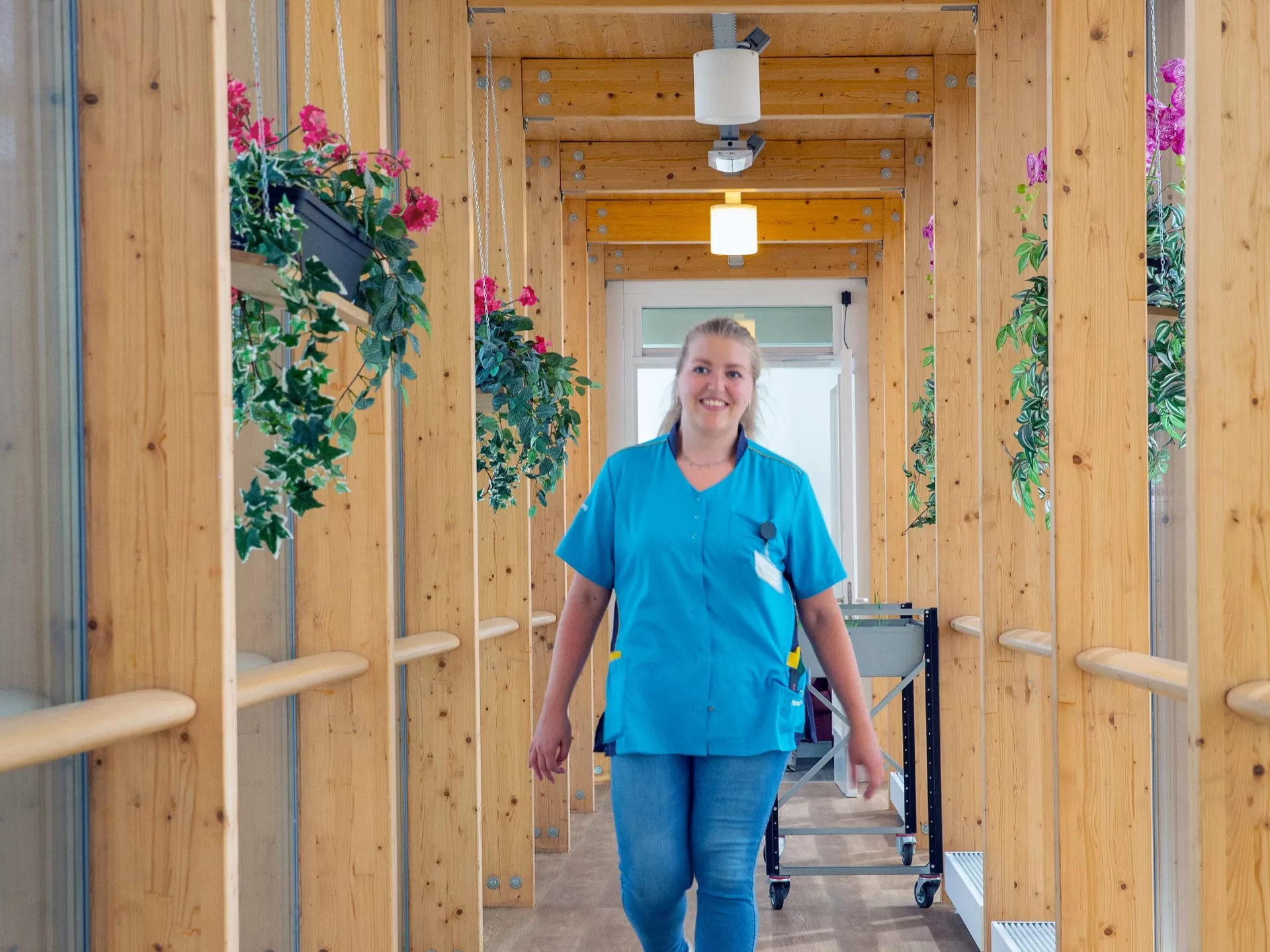 Een zorgverlener loopt door een houten gang met bloemen aan de zijkanten.