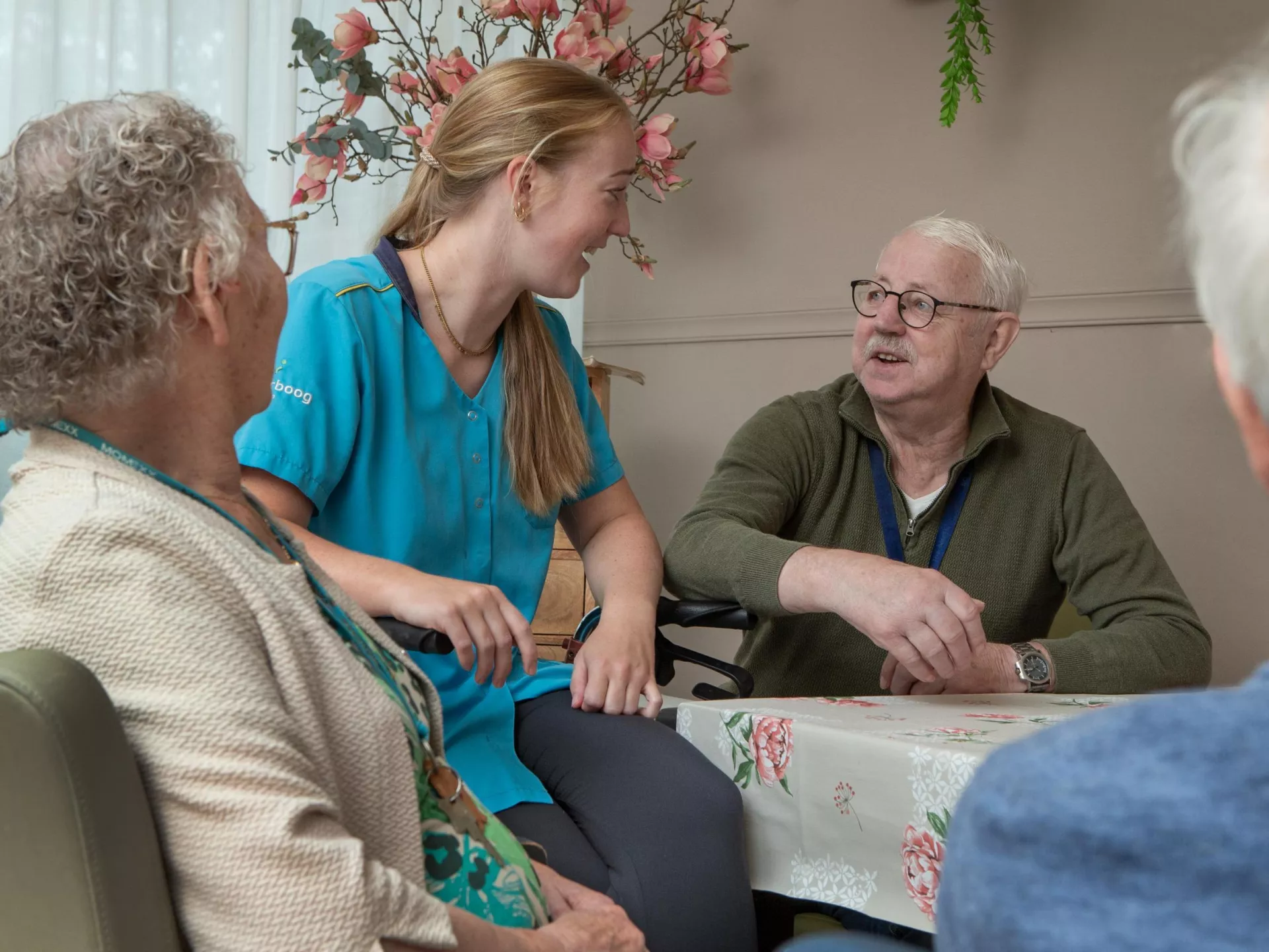 Verzorgster praat met senioren aan tafel in een huiselijke setting.