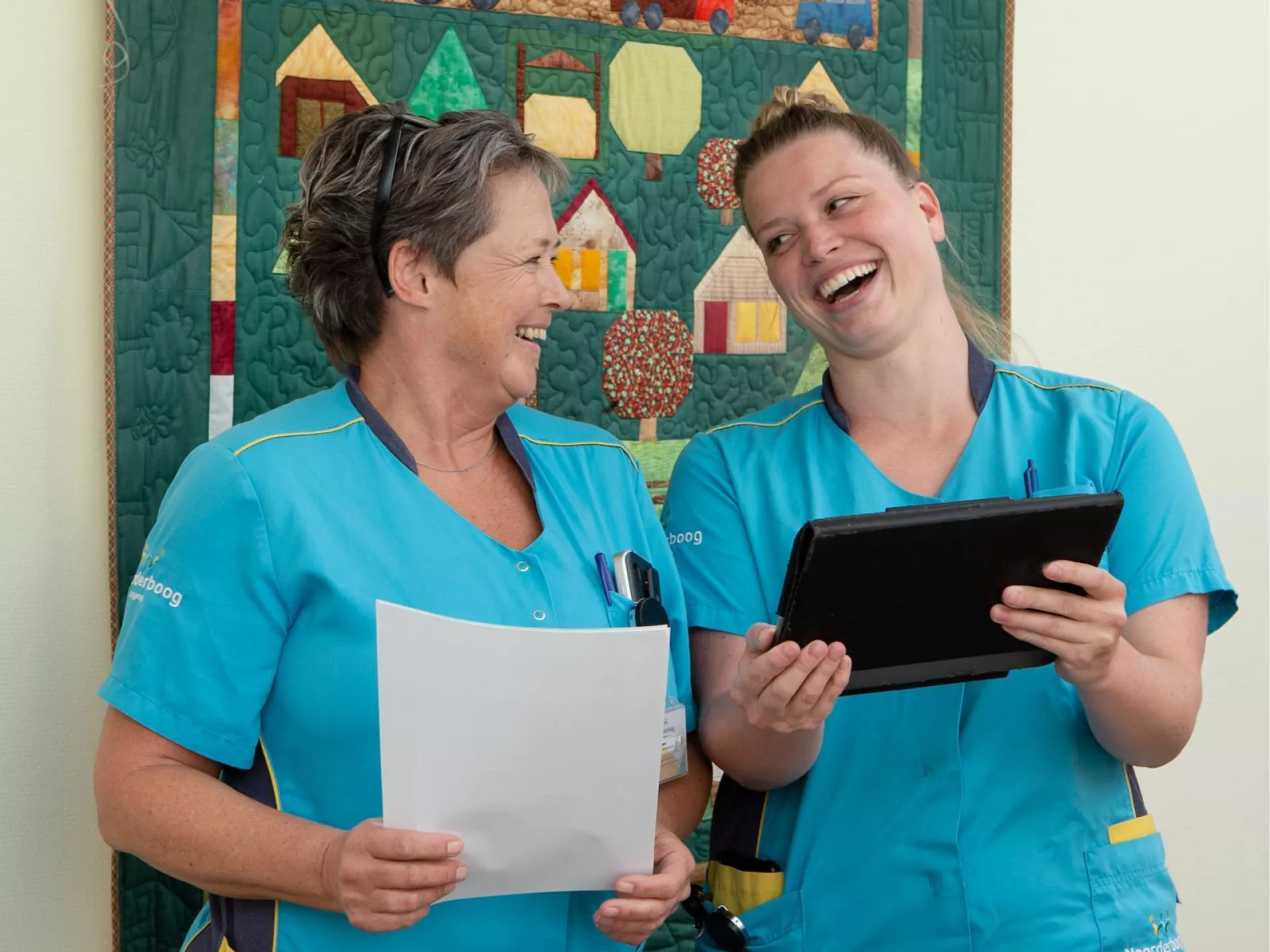 Twee verpleegkundigen lachen samen terwijl ze documenten en een tablet bekijken.