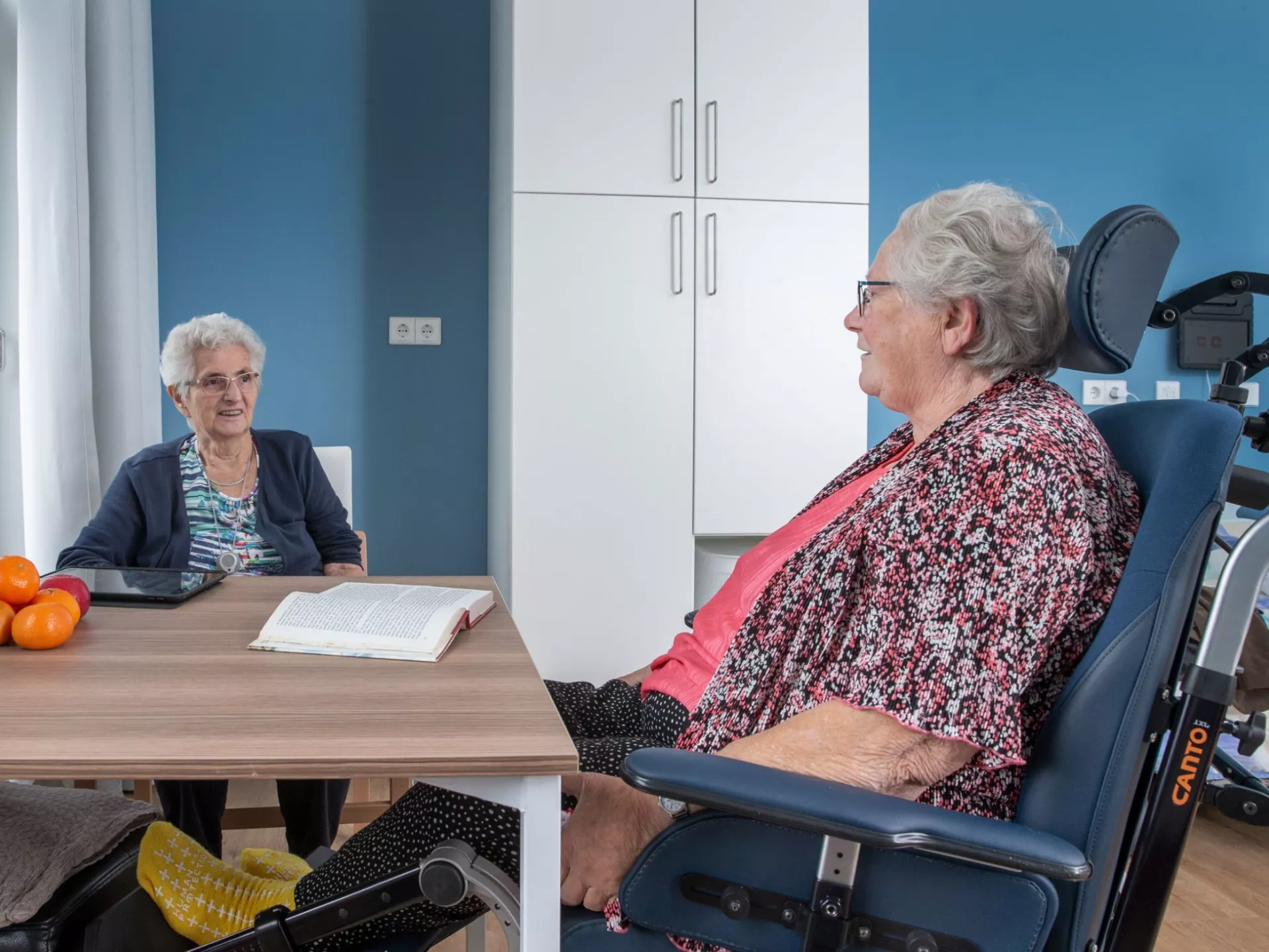 Twee oudere vrouwen zitten gezellig aan tafel in een kamer.