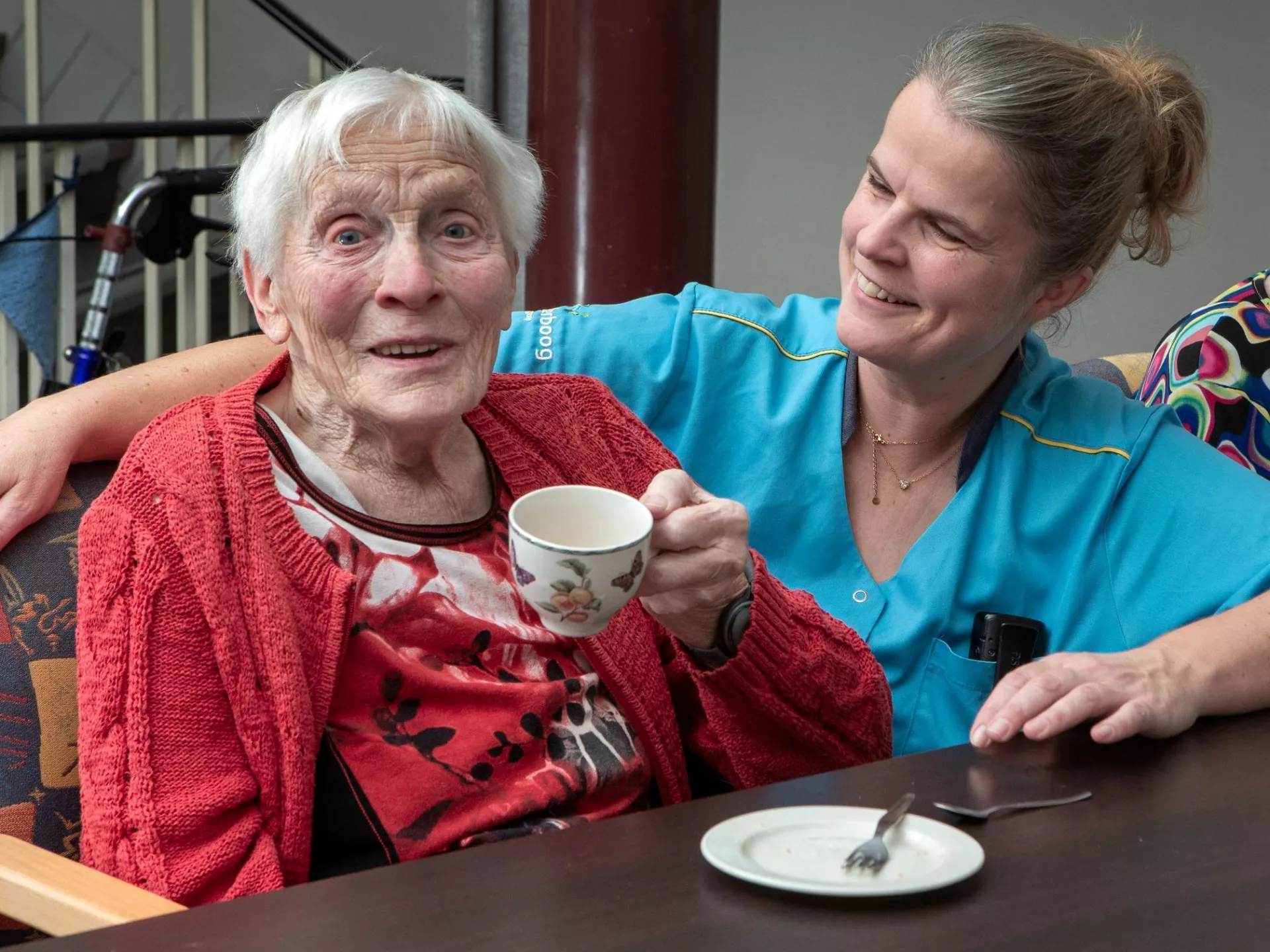Een oudere vrouw drinkt thee terwijl ze samen met een verzorgende lacht.