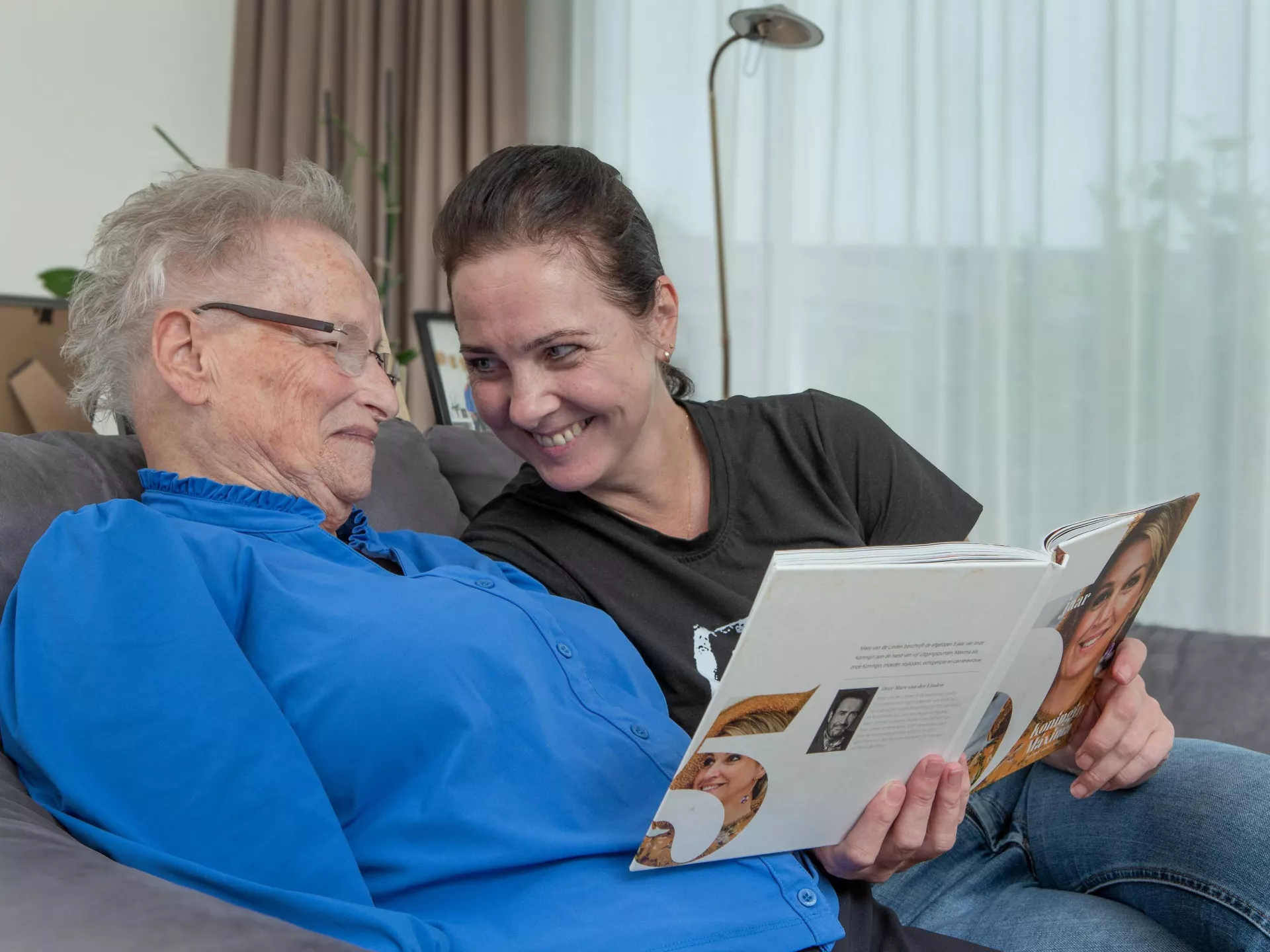 Een vrolijke vrouw leest een boek voor aan een oudere vrouw op de bank.