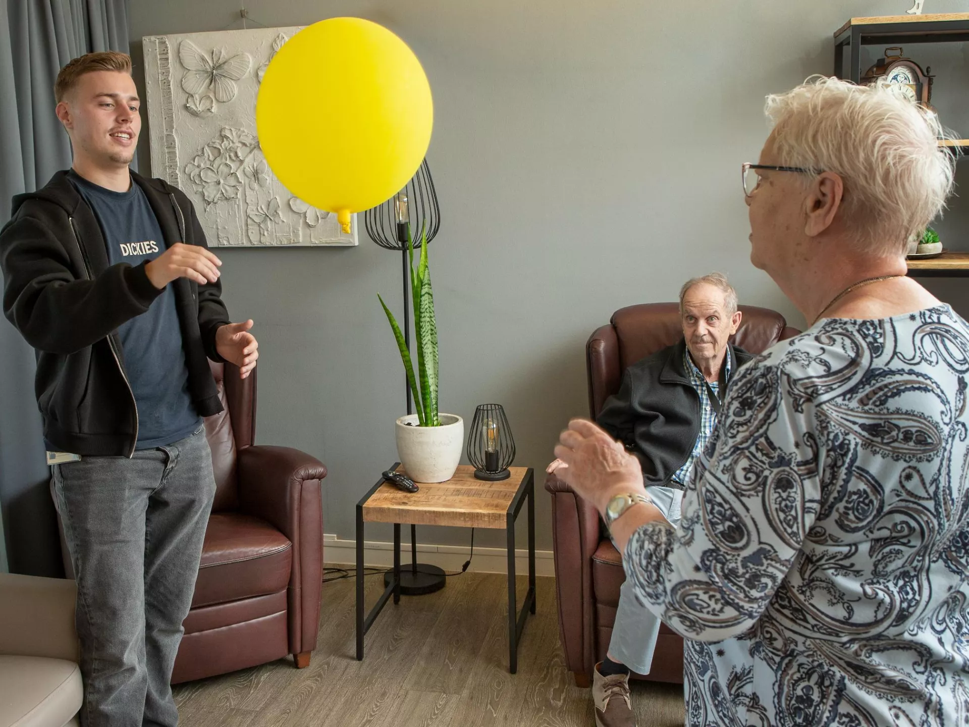 Een jonge man en een oudere vrouw spelen met een gele ballon in een kamer.