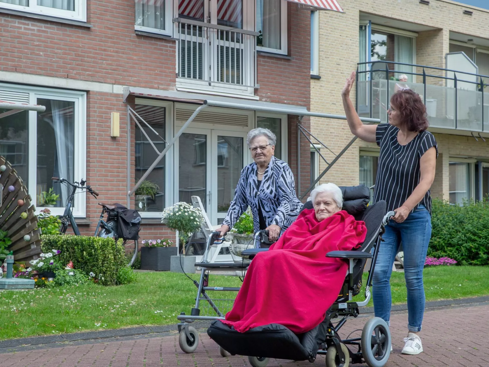 Twee vrouwen duwen een oudere vrouw in een rolstoel buiten.