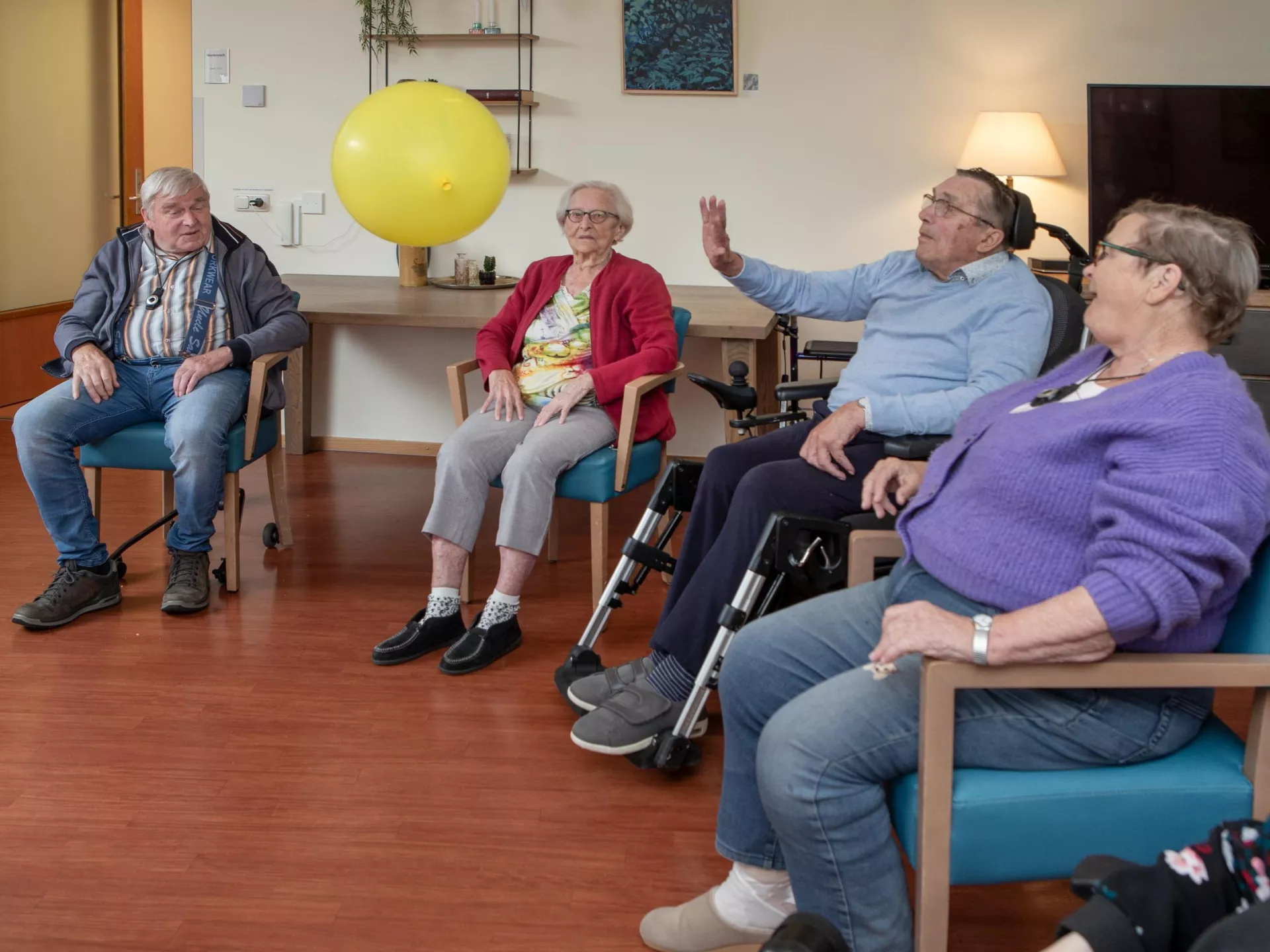 Een groep ouderen speelt een spel met een gele ballon in een gemeenschappelijke ruimte.