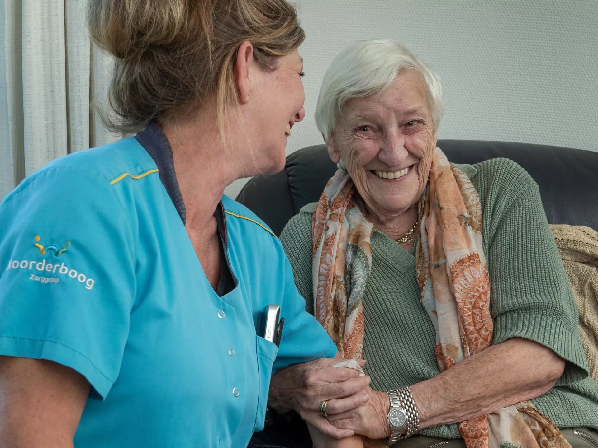 Een verzorgster en een oudere vrouw lachen samen op de bank.