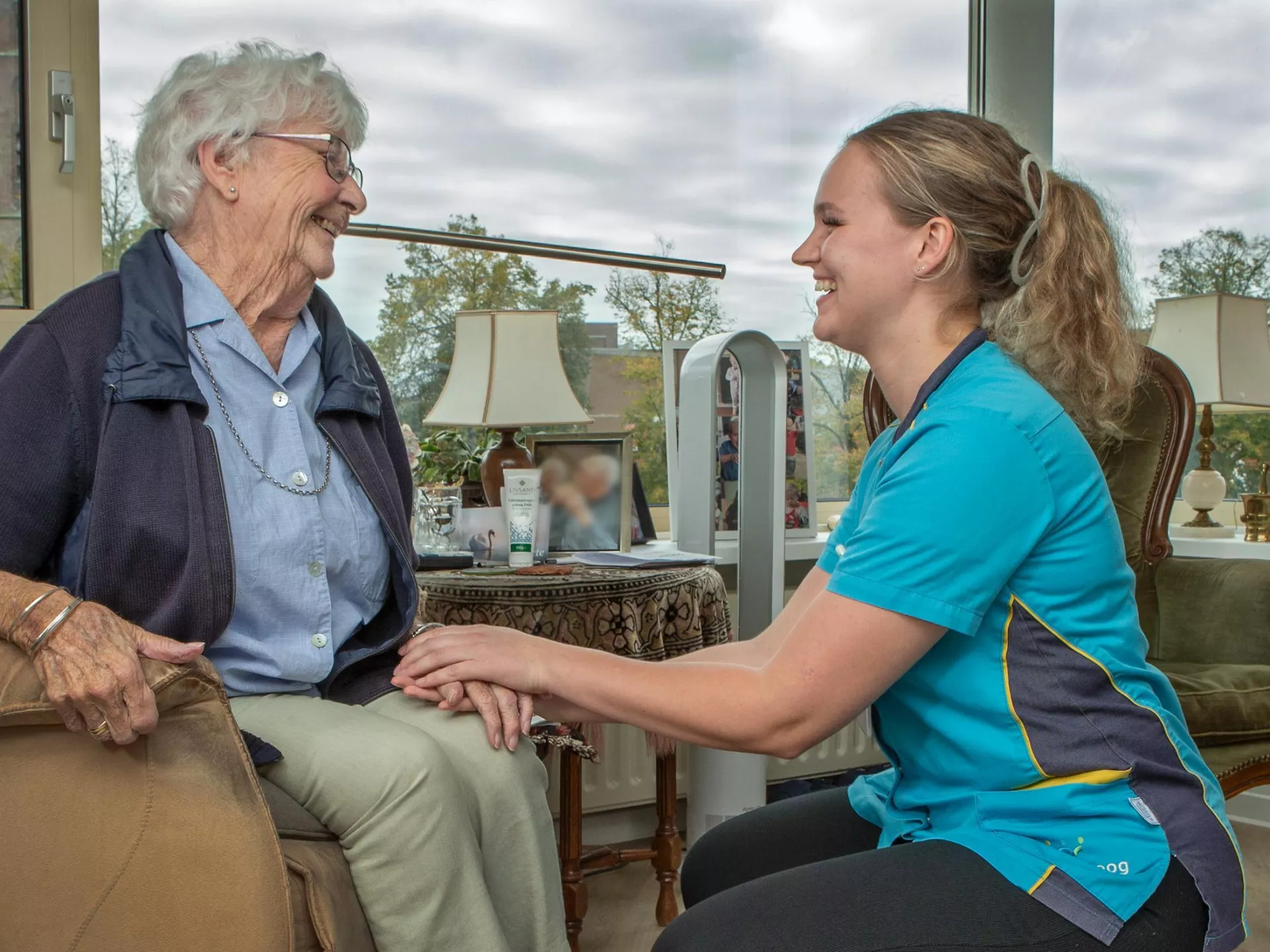 Een oudere vrouw glimlacht terwijl ze een verzorgster ontmoet in een gezellige kamer.