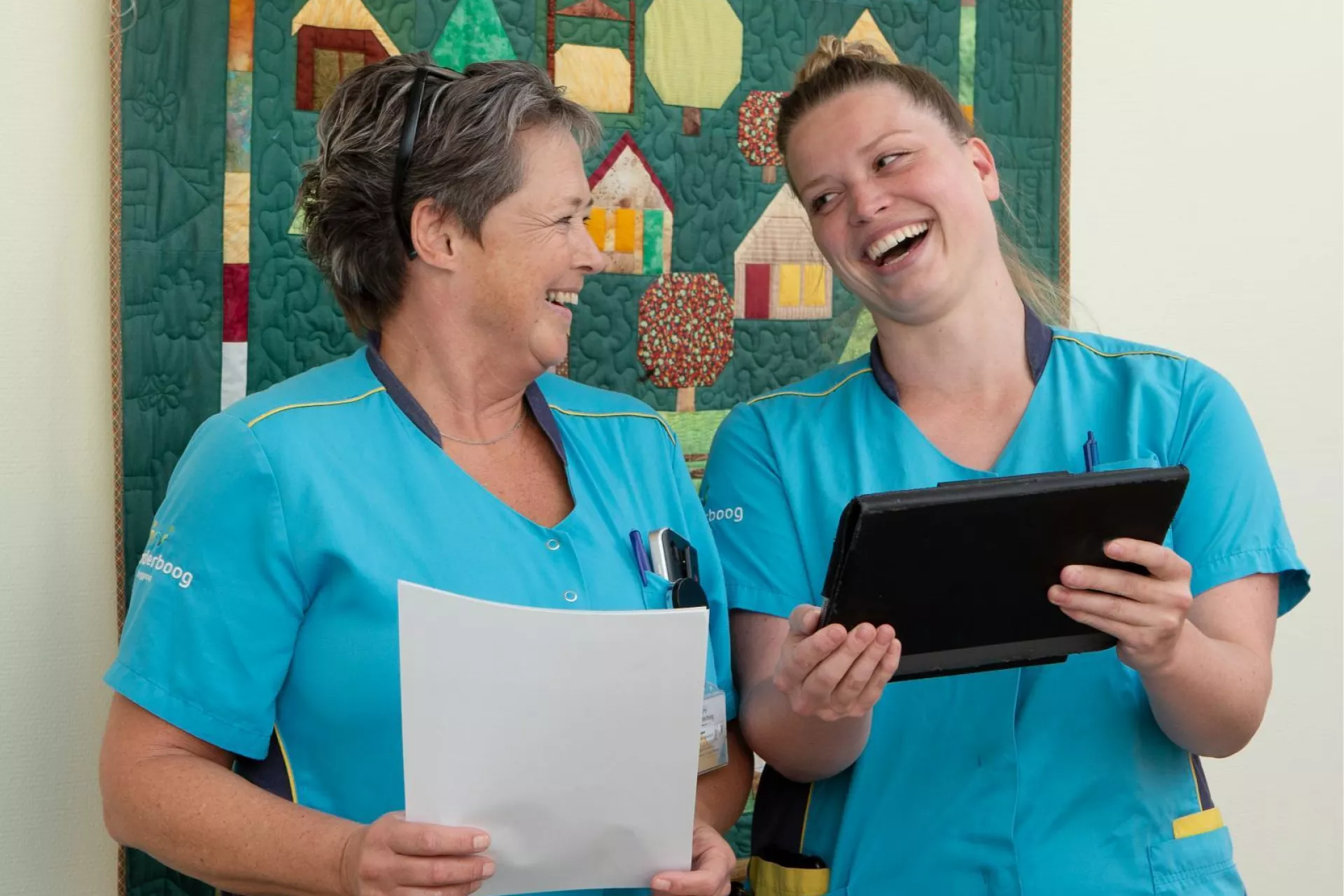 Twee verpleegkundigen lachen samen terwijl ze documenten en een tablet bekijken.